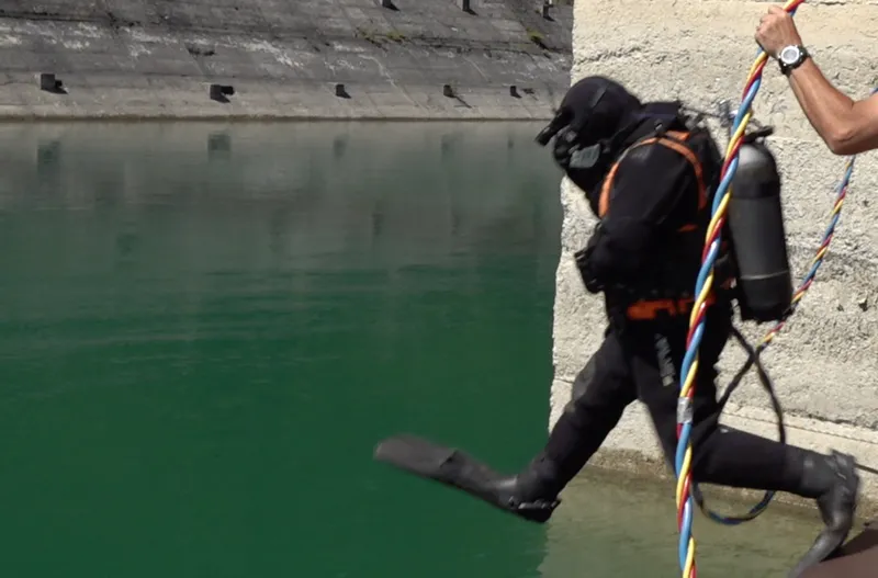 Scafandru cu furtun ombilical intrând în apă de pe ponton, lângă un perete de beton.