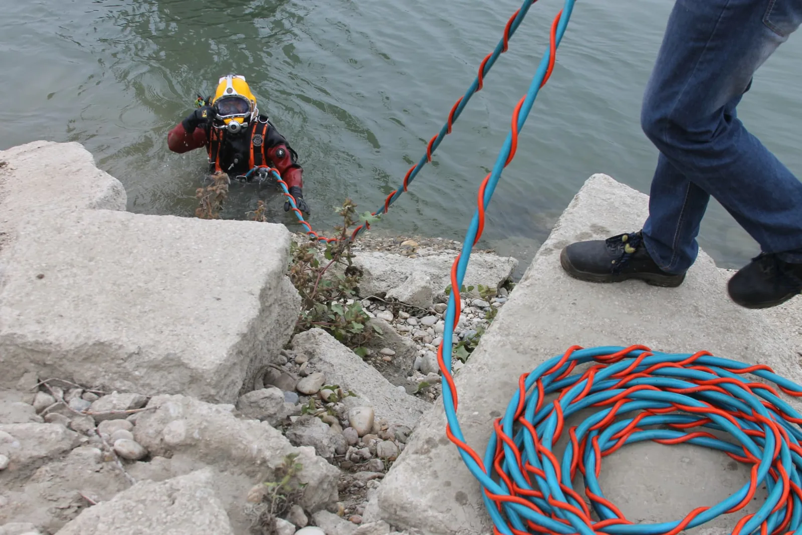 Scafandru echipat cu cască galbenă ieșind din apă pe malul unui lac de acumulare, cu furtunul ombilical desfășurat pe mal.