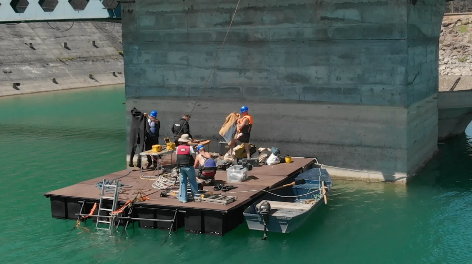 Ponton de lucru cu echipa de scafandri și echipamente, ancorat lângă pila de beton a unui turn de priză.