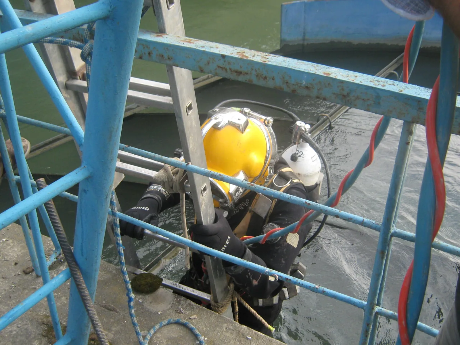 Scafandru comercial coborând pe scară către o structură metalică submersată, pregătit pentru lucrări de sudură subacvatică.
