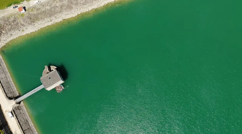 Vedere aeriană a unui turn de priză pe un lac de acumulare, cu pasarelă de acces și pontonul echipei de scafandri alături.